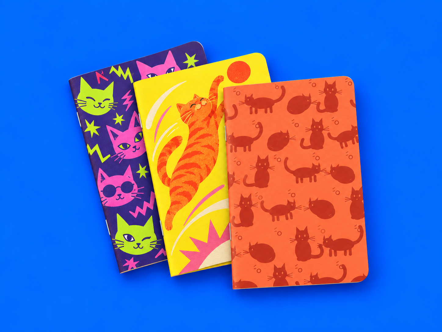 Tres libretas personalizadas con diseños de gatos en morado, amarillo y naranja sobre fondo azul